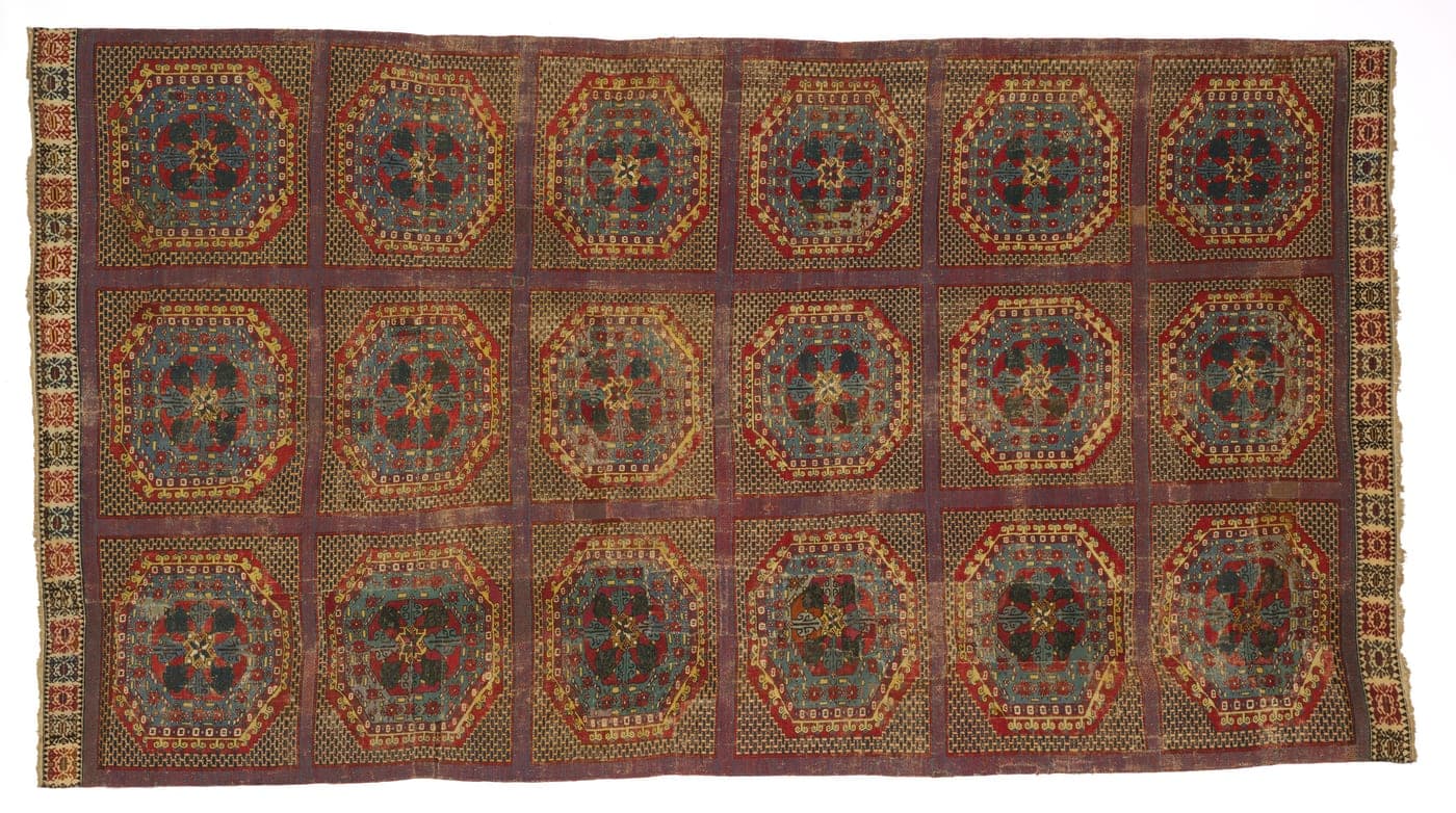 A late medieval Spanish carpet with a small-scale repeating geometric field in muted red, blue and gold, framed by wide patterned borders.