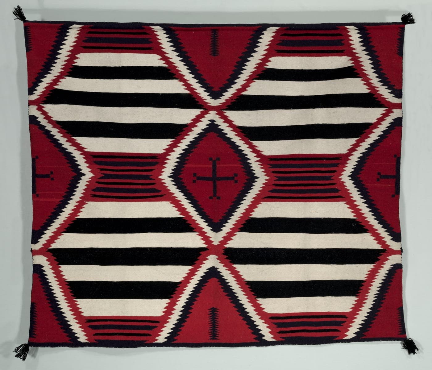 A Diné weaving in horizontal bands of red, black, white and grey, with stepped diamond forms breaking across the bands, wider than it is tall.