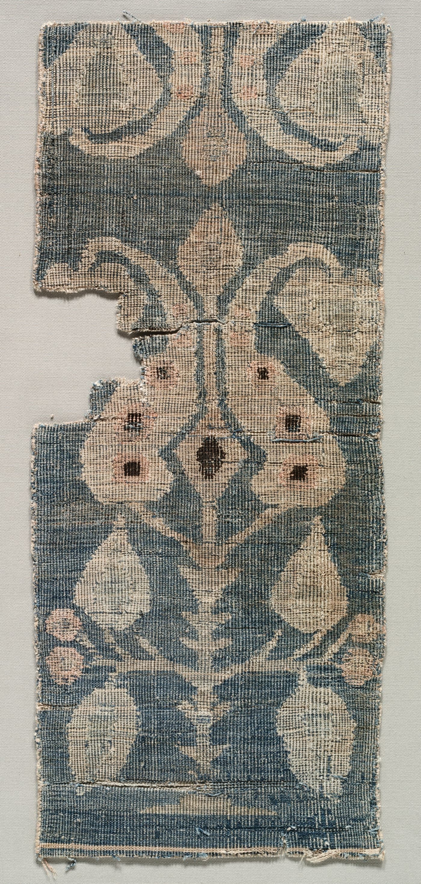 A worn carpet fragment in faded red and blue, showing part of a repeating geometric field and a fragment of border, the pile low and the foundation visible in places.